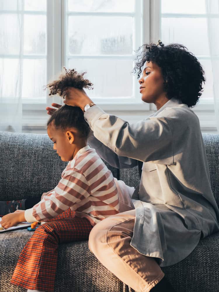 A woman sits behind her young daughter on a couch as she does her hair.