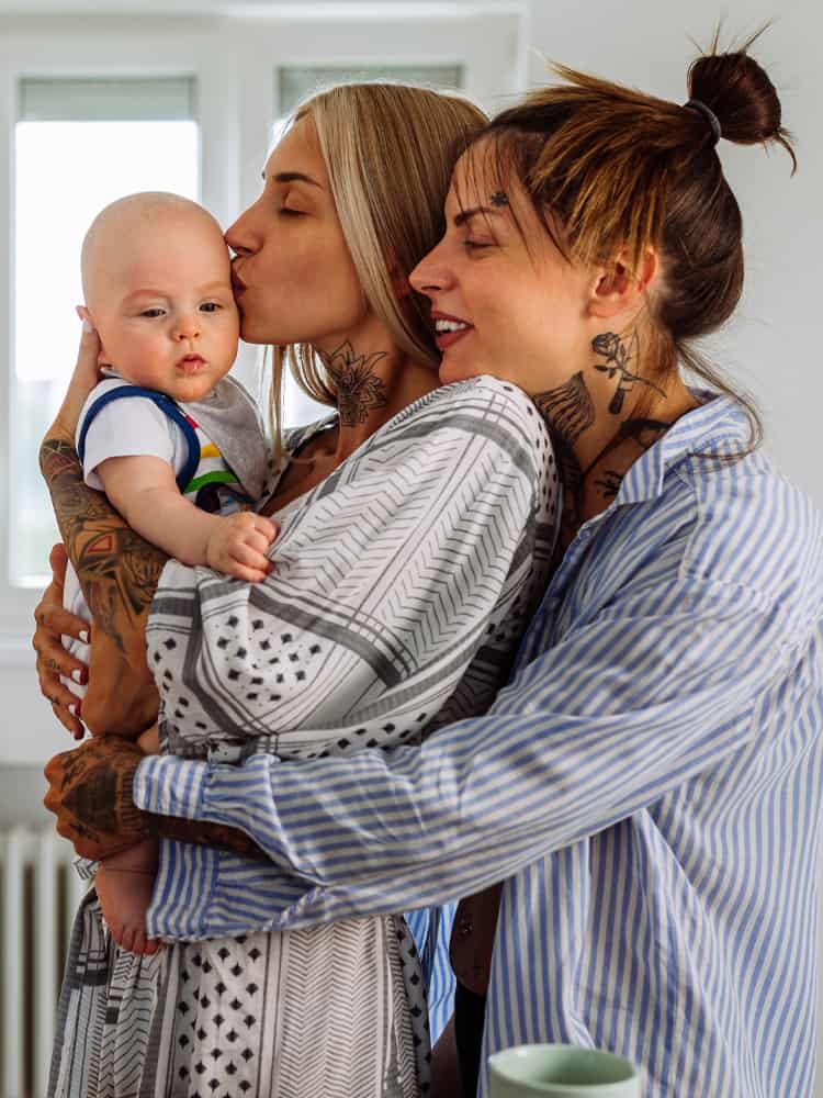 Woman holds and kisses a baby while another woman embraces her from behind.