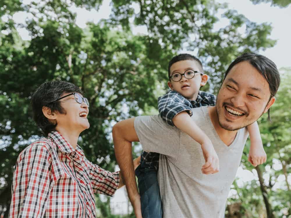 Man and woman laugh in a park while giving a child a piggy back ride.